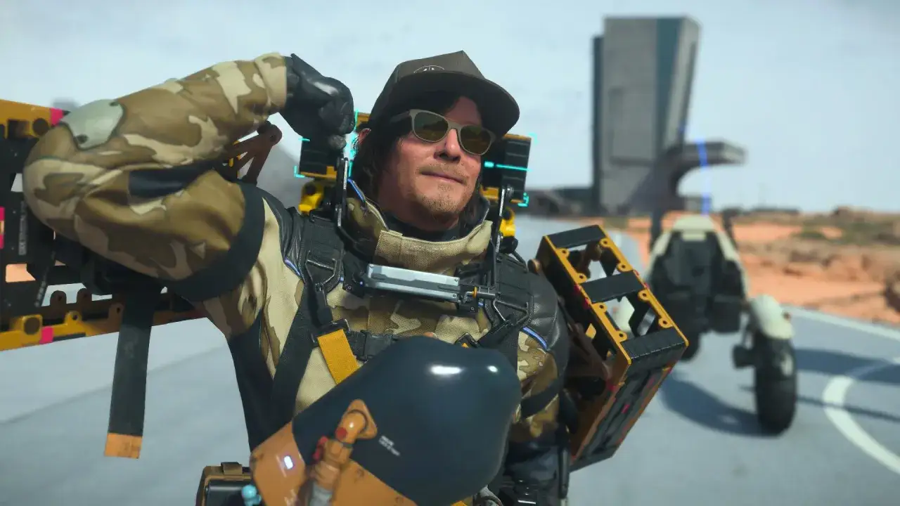 Sam Porter Bridges, con gafas de sol y gorra, listo para una nueva entrega en Death Stranding 2. Lleva su equipo de carga y un vehículo de fondo.